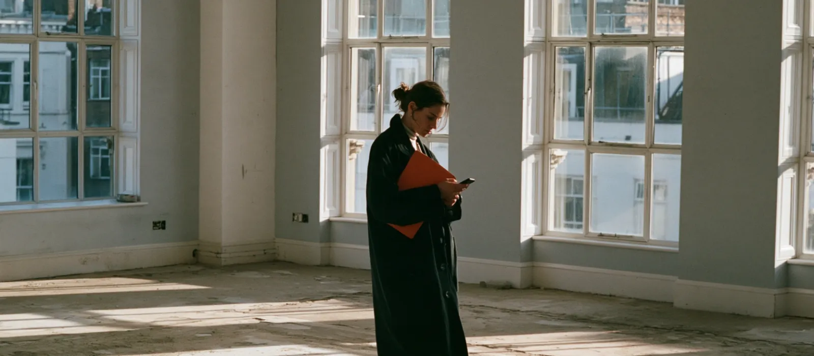 Estate agent in the empty living room of a flat, checking the listing record on her phone before a viewing