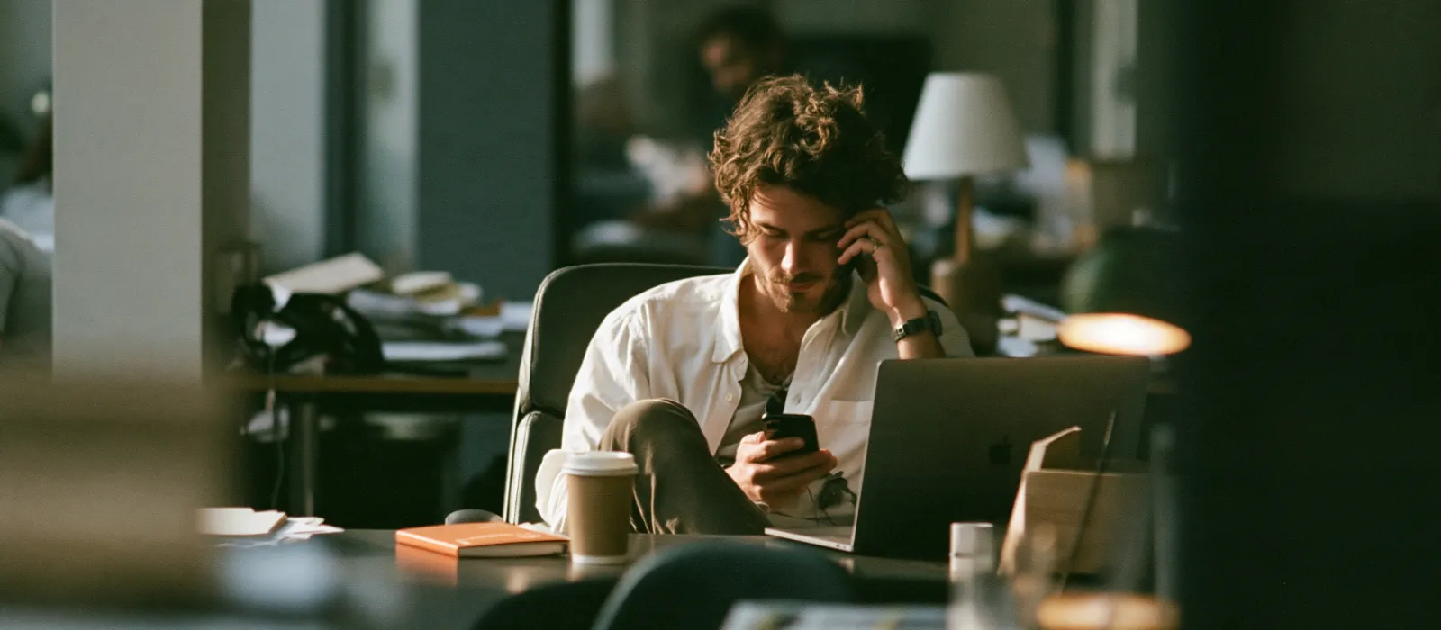 A young salesperson at his office desk in the morning replying to a message on his phone, an open laptop and a coffee beside him
