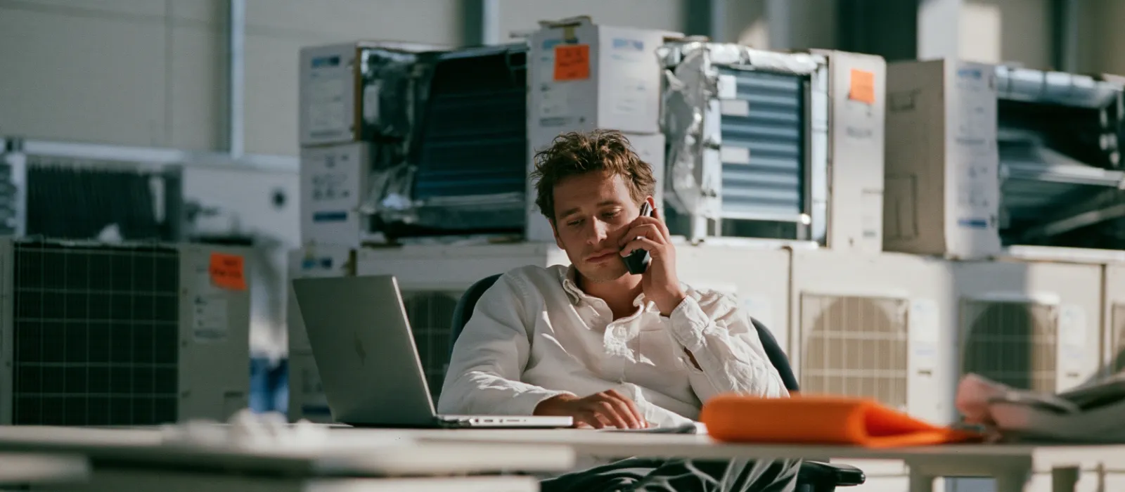Young employee at a desk in an air conditioning company office typing a message with a phone and laptop, boxed indoor units behind him